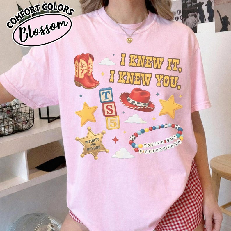 May include: A light pink t-shirt featuring a graphic print with the text I KNEW IT, I KNEW YOU, a red cowboy boot, a red cowboy hat, three alphabet blocks labeled T, S, 5, two yellow stars, a sheriff badge, and a colorful beaded bracelet.