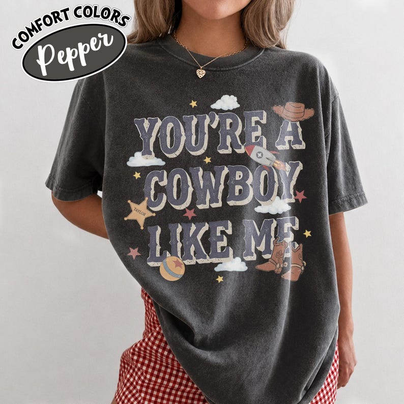 May include: A charcoal grey short-sleeve t-shirt featuring the text "YOU'RE A COWBOY LIKE ME" in a distressed font. The graphic includes illustrations of a rocket, a cowboy hat, boots, stars, clouds, and a ball, all in muted earth tones.