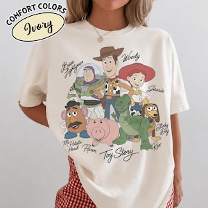 May include: Ivory-colored t-shirt featuring a graphic of Toy Story characters, including Woody, Buzz Lightyear, and Jessie. The shirt has the words "Toy Story" printed at the bottom. The shirt also has the words "COMFORT COLORS" and "Ivory" printed on the top left.