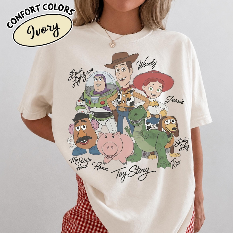 May include: Ivory-colored t-shirt featuring a graphic of Toy Story characters, including Woody, Buzz Lightyear, and Jessie. The shirt has the words "Toy Story" printed at the bottom. The shirt also has the words "COMFORT COLORS" and "Ivory" printed on the top left.
