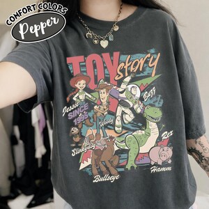 May include: Dark gray Comfort Colors t-shirt featuring a Toy Story graphic. The design includes characters like Woody, Buzz Lightyear, and Rex, with the text "Toy Story" and "Jessie Since 1995".