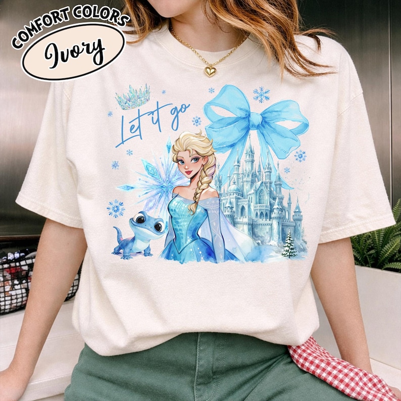 Puede incluir: Camiseta de color marfil con un gr&aacute;fico de Elsa de Frozen, un lazo azul y un castillo. El texto "Let it go" est&aacute; escrito en azul. La camiseta tiene mangas cortas y cuello redondo. Las palabras "Comfort Colors" e "Ivory" est&aacute;n en un c&iacute;rculo en la parte superior izquierda.