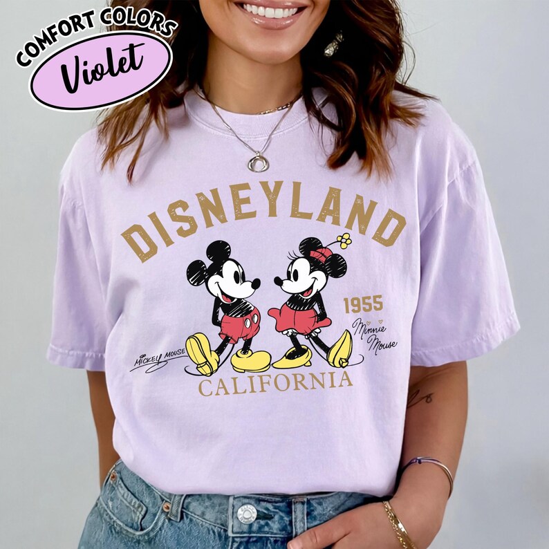 K&ouml;nnte beinhalten: Ein violettes Comfort Colors T-Shirt mit einem Disneyland-Motiv. Das Design zeigt Micky und Minnie Maus, den Text "Disneyland 1955 California" und die Unterschriften "Mickey Mouse" und "Minnie Mouse".