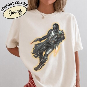 Puede incluir: Una camiseta de color marfil con un gr&aacute;fico de un personaje con armadura plateada que sostiene a una peque&ntilde;a criatura verde. El personaje lleva casco y capa, y sostiene un bl&aacute;ster. El texto en la esquina superior izquierda dice COMFORT COLORS Ivory.