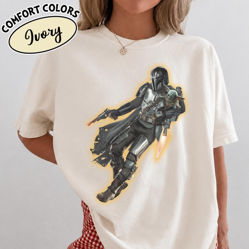 Puede incluir: Una camiseta de color marfil con un gr&aacute;fico de un personaje con armadura plateada que sostiene a una peque&ntilde;a criatura verde. El personaje lleva casco y capa, y sostiene un bl&aacute;ster. El texto en la esquina superior izquierda dice COMFORT COLORS Ivory.