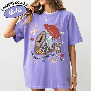 Puede incluir: Una camiseta violeta de manga corta con un estampado gr&aacute;fico de una bota de vaquero con manchas de vaca, un sombrero de vaquero rojo, un lazo y bloques de letras. El texto "NAME'S" y "I KNEW IT I KNEW YOU" aparece junto a iconos de estrellas y flores.