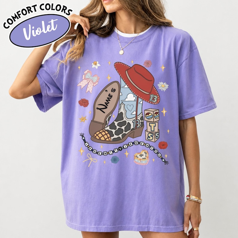 Puede incluir: Una camiseta violeta de manga corta con un estampado gr&aacute;fico de una bota de vaquero con manchas de vaca, un sombrero de vaquero rojo, un lazo y bloques de letras. El texto "NAME'S" y "I KNEW IT I KNEW YOU" aparece junto a iconos de estrellas y flores.