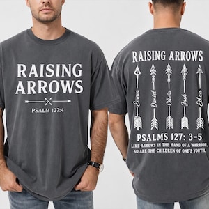 Op de afbeelding: Een antracietgrijs t-shirt met korte mouwen en witte tekst. De voorkant toont "RAISING ARROWS" en "PSALM 127:4". De achterkant toont vijf verticale pijlen met namen, "PSALMS 127: 3-5", en de tekst "LIKE ARROWS IN THE HAND OF A WARRIOR, SO ARE THE CHILDREN OF ONE'S YOUTH."