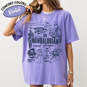Op de afbeelding: Een violet Comfort Colors T-shirt met een grafisch ontwerp van personages en elementen uit de serie The Mandalorian. Het ontwerp bevat de tekst "The Mandalorian and Grogu" en "This is the Way."