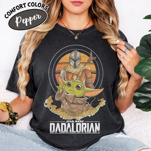 Może przedstawiać: Czarna koszulka z grafiką przedstawiającą The Mandalorian i Grogu z Gwiezdnych Wojen. Projekt zawiera okrągły obraz postaci z tekstem "STAR WARS DADALORIAN" poniżej. Koszulka ma tekst "COMFORT COLORS Pepper" w owalu.