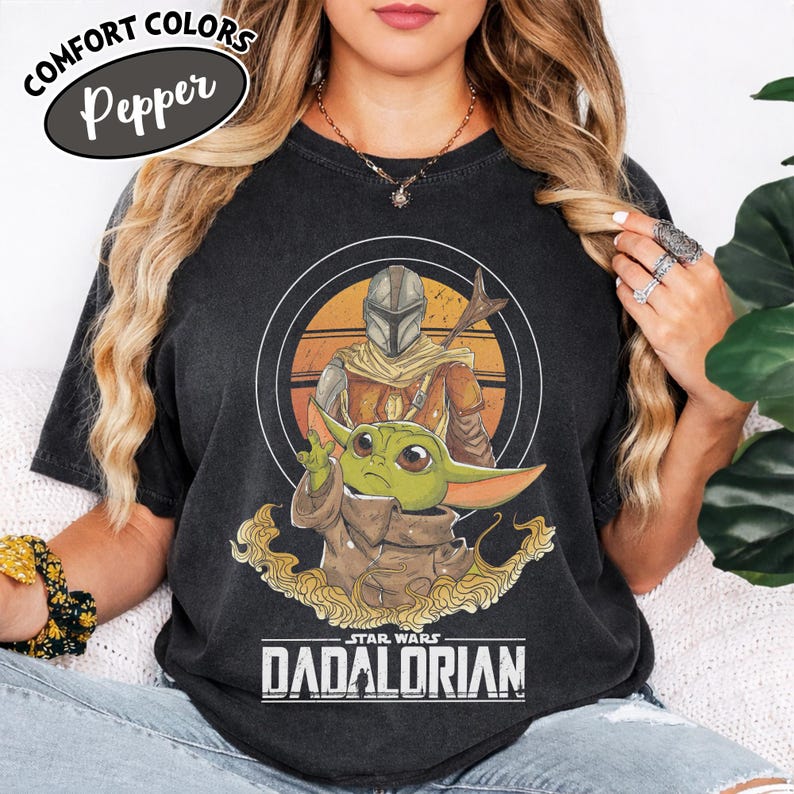 Może przedstawiać: Czarna koszulka z grafiką przedstawiającą The Mandalorian i Grogu z Gwiezdnych Wojen. Projekt zawiera okrągły obraz postaci z tekstem "STAR WARS DADALORIAN" poniżej. Koszulka ma tekst "COMFORT COLORS Pepper" w owalu.