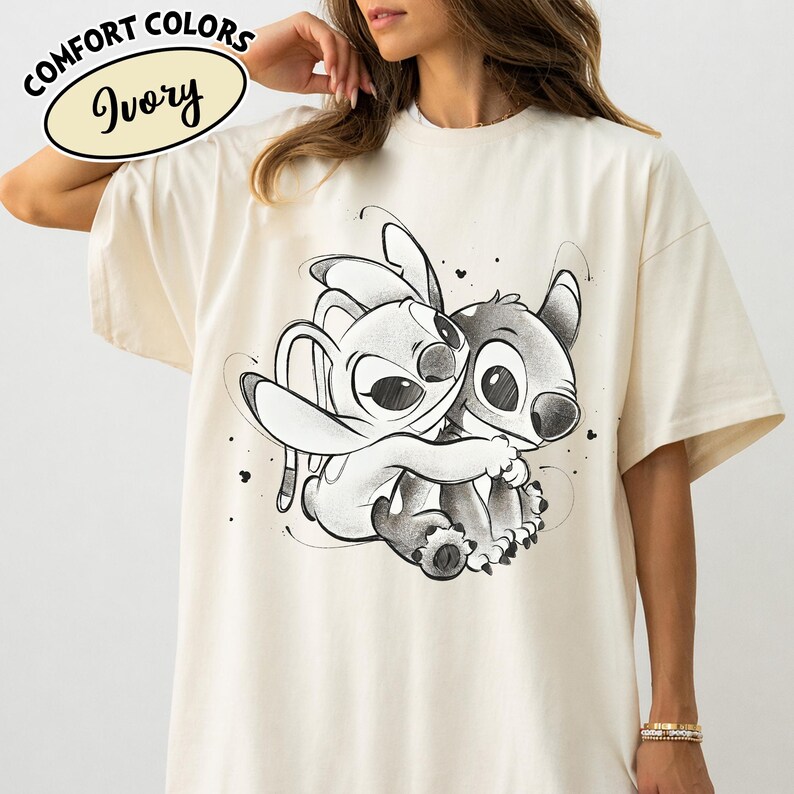May include: Ivory-colored t-shirt featuring a black and white illustration of two cartoon characters embracing. The shirt has the words "Comfort Colors" and "Ivory" printed on it.