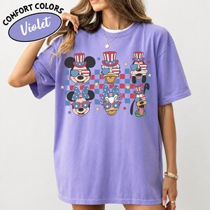 May include: Violet Comfort Colors t-shirt featuring cartoon characters in patriotic attire. Mickey Mouse, Donald Duck, Goofy, Minnie Mouse, and Daisy Duck are depicted wearing sunglasses and hats with American flag designs. The shirt has a checkered background.