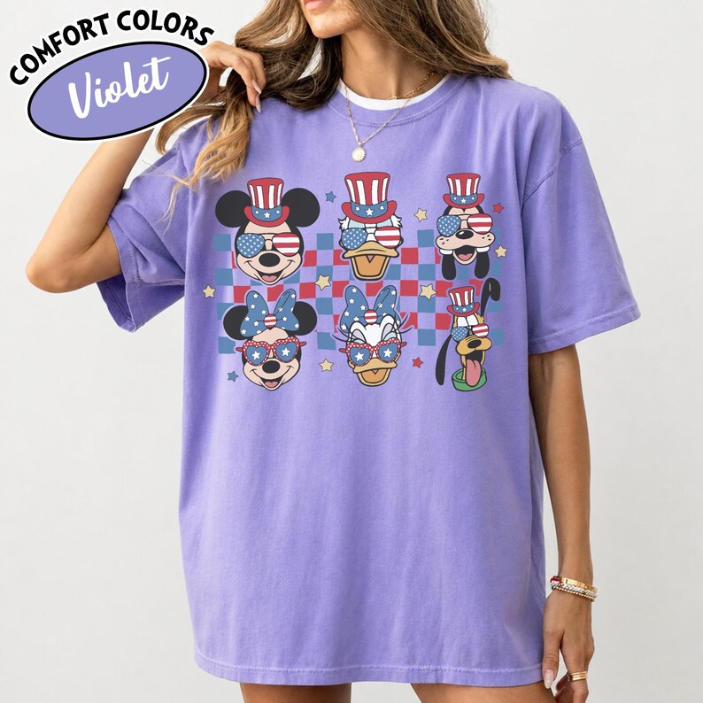 May include: Violet Comfort Colors t-shirt featuring cartoon characters in patriotic attire. Mickey Mouse, Donald Duck, Goofy, Minnie Mouse, and Daisy Duck are depicted wearing sunglasses and hats with American flag designs. The shirt has a checkered background.