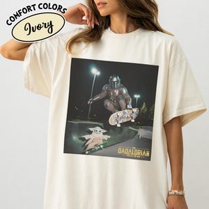 May include: Ivory-colored t-shirt featuring a graphic of The Mandalorian and Grogu skateboarding in a skate park. The graphic includes the text "THE DADALORIAN" and "THIS IS THE WAY". The shirt has the words "COMFORT COLORS" and "Ivory" printed on the top left.