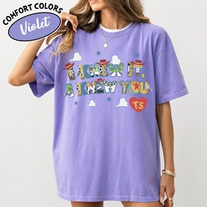 May include: A violet short-sleeve t-shirt featuring a graphic print with the text I KNEW IT, I KNEW YOU and a red heart with the letters TS. The letters are decorated with patterns inspired by cowboy and space ranger characters, including stars and clouds.