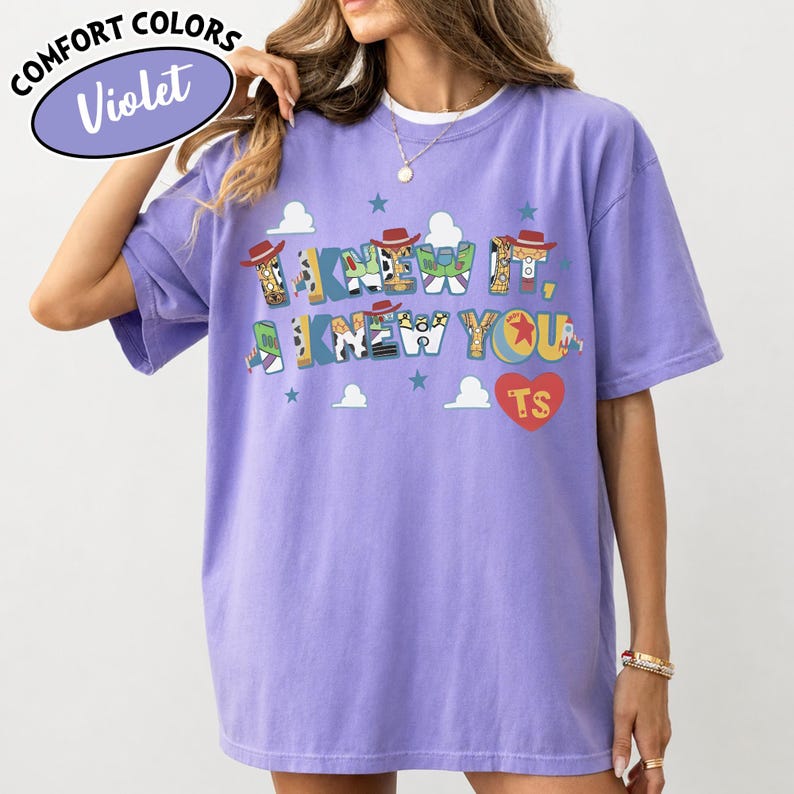 May include: A violet short-sleeve t-shirt featuring a graphic print with the text I KNEW IT, I KNEW YOU and a red heart with the letters TS. The letters are decorated with patterns inspired by cowboy and space ranger characters, including stars and clouds.