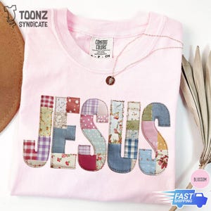 May include: A light pink t-shirt with the word "JESUS" appliquéd across the front in a patchwork design. The letters are made of various patterned fabrics. A gold necklace with a pendant rests on the shirt.