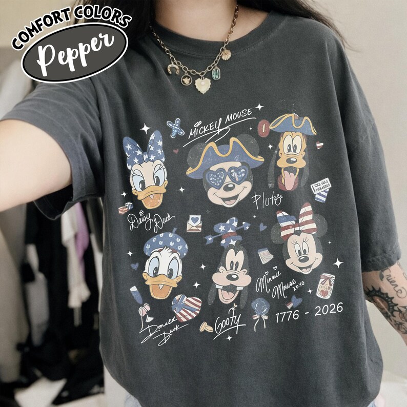 May include: A charcoal grey short-sleeve t-shirt featuring a graphic print of Disney characters including Mickey Mouse, Minnie Mouse, Donald Duck, Daisy Duck, Goofy, and Pluto. The design includes patriotic stars and stripes motifs and the text 1776 - 2026.