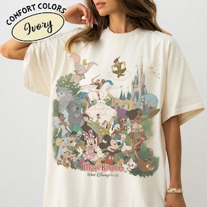 Op de afbeelding: Ivoor kleurig T-shirt met een kleurrijke afbeelding van verschillende Disney-personages, waaronder Mickey Mouse, Dumbo en Peter Pan, met de woorden "Magic Kingdom" en "Walt Disney World" eronder gedrukt. Het shirt heeft ook de woorden "Comfort Colors" en "Ivory" linksboven gedrukt.