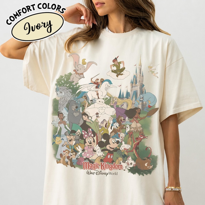 Op de afbeelding: Ivoor kleurig T-shirt met een kleurrijke afbeelding van verschillende Disney-personages, waaronder Mickey Mouse, Dumbo en Peter Pan, met de woorden "Magic Kingdom" en "Walt Disney World" eronder gedrukt. Het shirt heeft ook de woorden "Comfort Colors" en "Ivory" linksboven gedrukt.