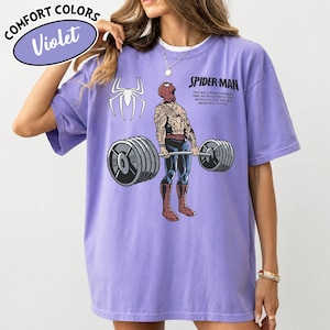 May include: Violet Comfort Colors t-shirt featuring a Spider-Man graphic lifting weights. The shirt includes the text "SPIDER-MAN" and a Spider-Man logo. The shirt is a light purple color.
