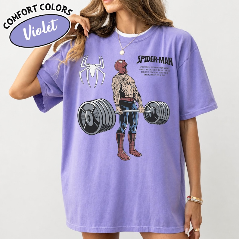 May include: Violet Comfort Colors t-shirt featuring a Spider-Man graphic lifting weights. The shirt includes the text "SPIDER-MAN" and a Spider-Man logo. The shirt is a light purple color.