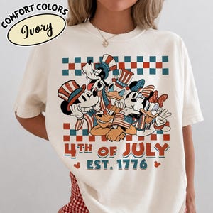 May include: An ivory t-shirt featuring a graphic of cartoon characters wearing patriotic hats and holding American flags. Above and below the characters are red and teal checkered patterns. The text reads 4TH OF JULY EST. 1776.