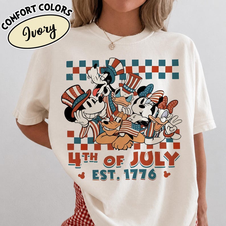 May include: An ivory t-shirt featuring a graphic of cartoon characters wearing patriotic hats and holding American flags. Above and below the characters are red and teal checkered patterns. The text reads 4TH OF JULY EST. 1776.