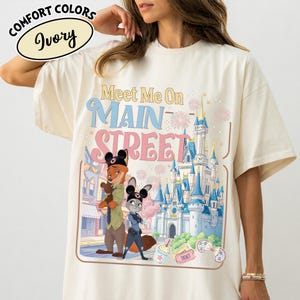 May include: Ivory-colored t-shirt featuring the text "Meet Me On Main Street" with cartoon characters and a castle illustration. The shirt has a relaxed fit and the words "Comfort Colors" are printed at the top.