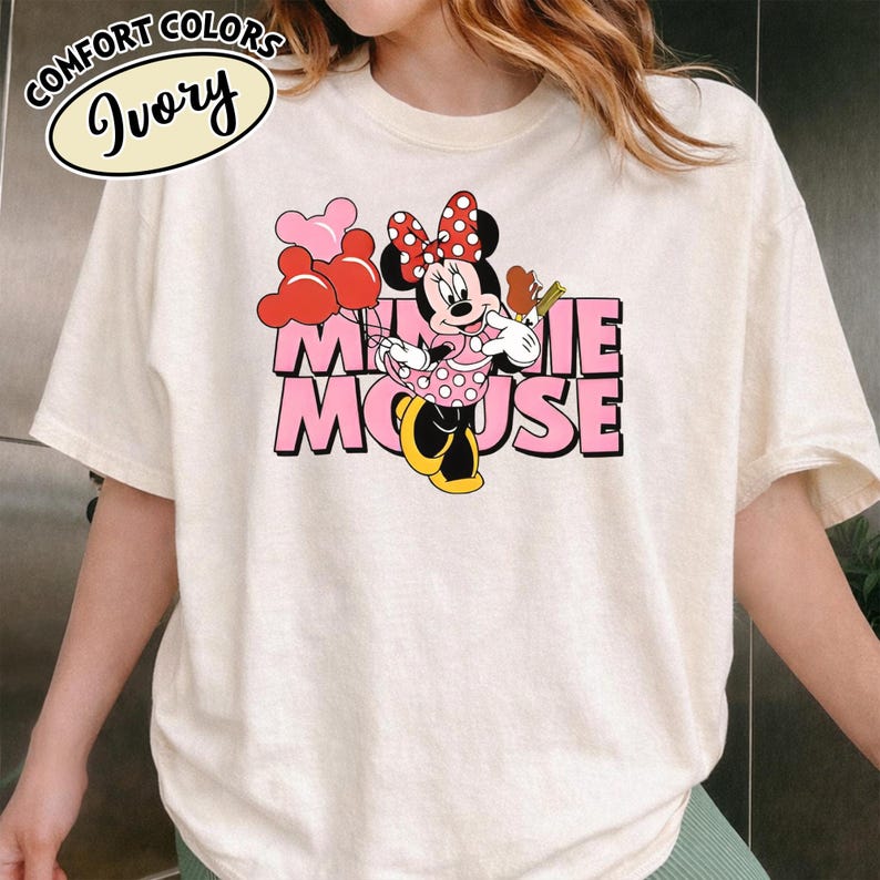 May include: Ivory t-shirt with a Minnie Mouse graphic. The design shows Minnie Mouse with balloons and a snack, and the words "Minnie Mouse" in pink. The shirt also has the words "Comfort Colors" and "Ivory" in a circular design.