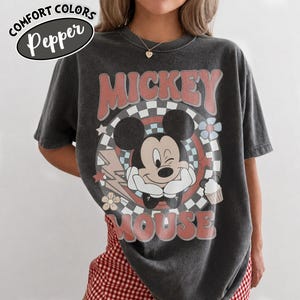 May include: Dark gray t-shirt featuring a graphic of Mickey Mouse winking, surrounded by a retro design with the words "Mickey Mouse." The shirt has the text "Comfort Colors Pepper" in a white oval on the top left.