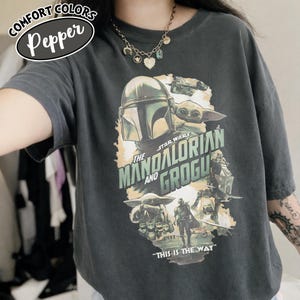 May include: Dark grey t-shirt featuring a graphic of The Mandalorian and Grogu from Star Wars. The design includes the text "THE MANDALORIAN AND GROGU" and "THIS IS THE WAY". The shirt has the Comfort Colors brand label.