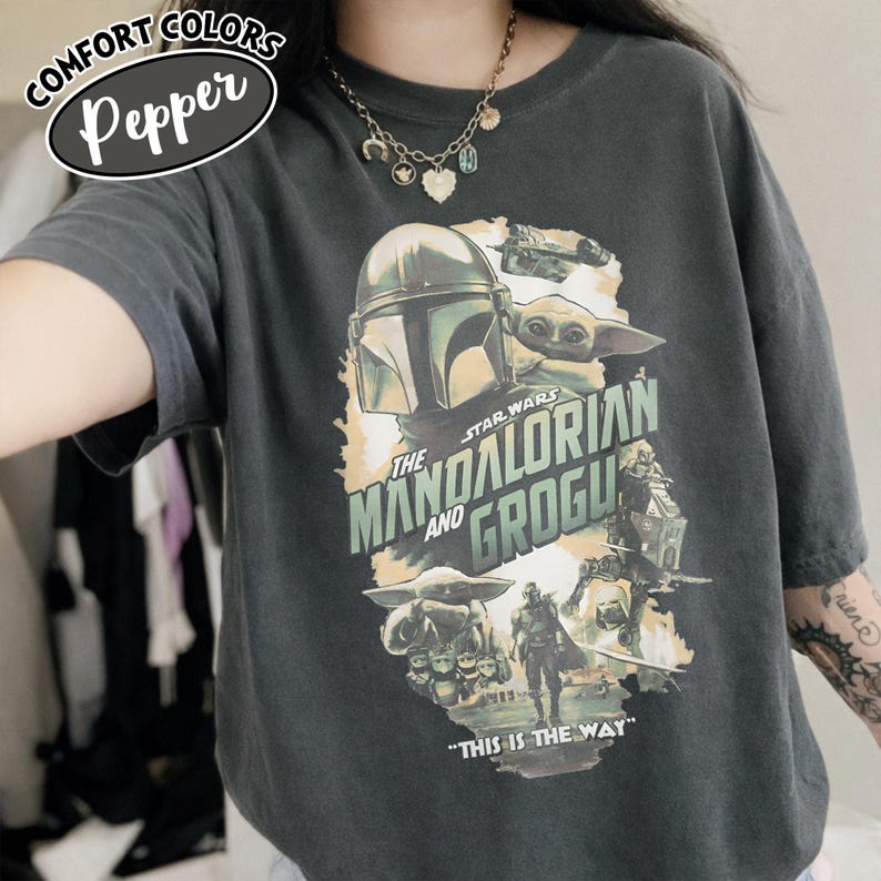May include: Dark grey t-shirt featuring a graphic of The Mandalorian and Grogu from Star Wars. The design includes the text "THE MANDALORIAN AND GROGU" and "THIS IS THE WAY". The shirt has the Comfort Colors brand label.