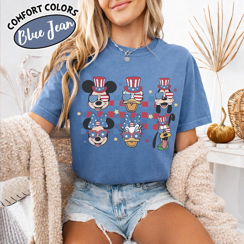 May include: Blue jean-colored t-shirt featuring cartoon characters with patriotic designs. The characters wear sunglasses, hats, and bows in red, white, and blue. The shirt has the words "Comfort Colors" and "Blue Jean" in a circular design.