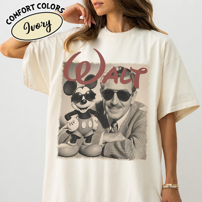 May include: Ivory-colored t-shirt featuring a sepia-toned image of Walt Disney and Mickey Mouse wearing sunglasses. The word "WALT" is printed above the image in a maroon color. The shirt has the words "COMFORT COLORS" and "Ivory" printed on the top left.