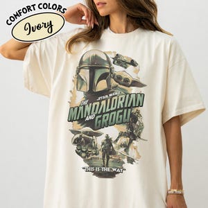 May include: Ivory-colored t-shirt featuring a graphic from the Star Wars series, "The Mandalorian and Grogu." The design includes images of the Mandalorian, Grogu, and other characters, with the phrase "This is the Way" at the bottom. The shirt has short sleeves.
