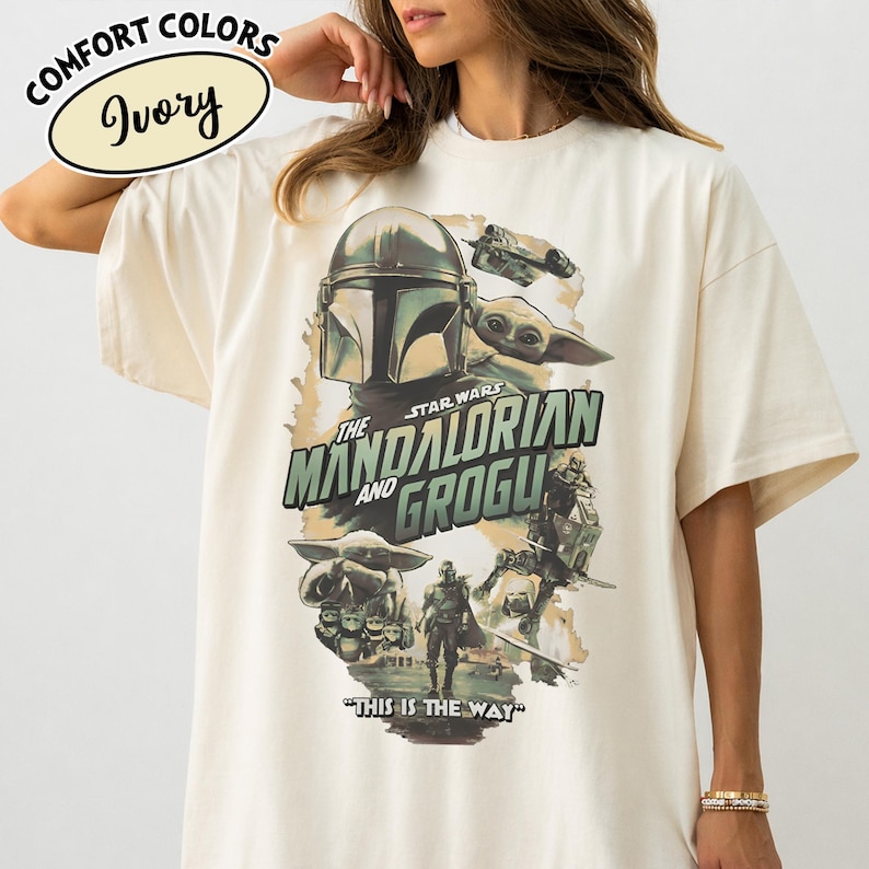 May include: Ivory-colored t-shirt featuring a graphic from the Star Wars series, "The Mandalorian and Grogu." The design includes images of the Mandalorian, Grogu, and other characters, with the phrase "This is the Way" at the bottom. The shirt has short sleeves.