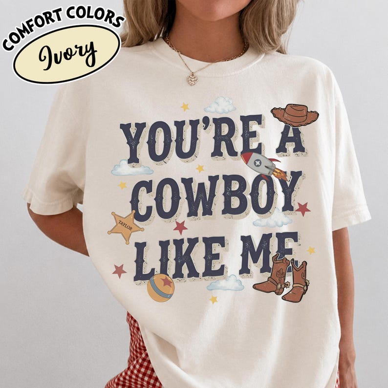May include: An ivory short-sleeve t-shirt featuring the dark blue text 'YOU'RE A COWBOY LIKE ME' with graphic illustrations of a brown cowboy hat, a rocket ship, a sheriff star, a ball, and cowboy boots, accented by small stars and clouds.