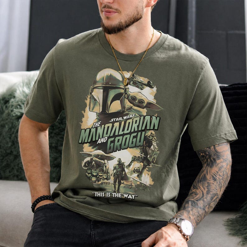 May include: Olive green t-shirt featuring a graphic from "The Mandalorian" series. The design includes the Mandalorian helmet, Grogu, and the text "The Mandalorian and Grogu" and "This is the Way".