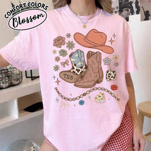May include: A light pink t-shirt featuring a graphic print of a tan cowboy hat, two western boots, flowers, stars, and a heart-shaped cake. The text TS5 is on one boot, and the text I KNEW IT Y I KNEW YOU is on a beaded string graphic.