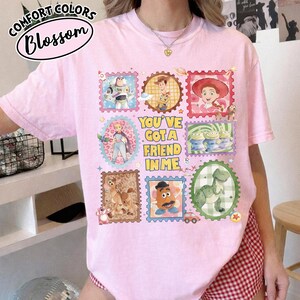 May include: Pink t-shirt with a colorful graphic featuring various characters from a popular animated movie. The design includes postage stamp-style images and the text "YOU'VE GOT A FRIEND IN ME". The shirt has the text "COMFORT COLORS Blossom" on the top left.