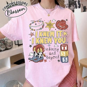 May include: A light pink t-shirt featuring a graphic print with the text I KNEW IT I KNEW YOU infinity and beyond. The design includes a beaded bracelet, a yellow sheriff star, a brown cowboy hat, cow-print boots, and three stacked alphabet blocks.