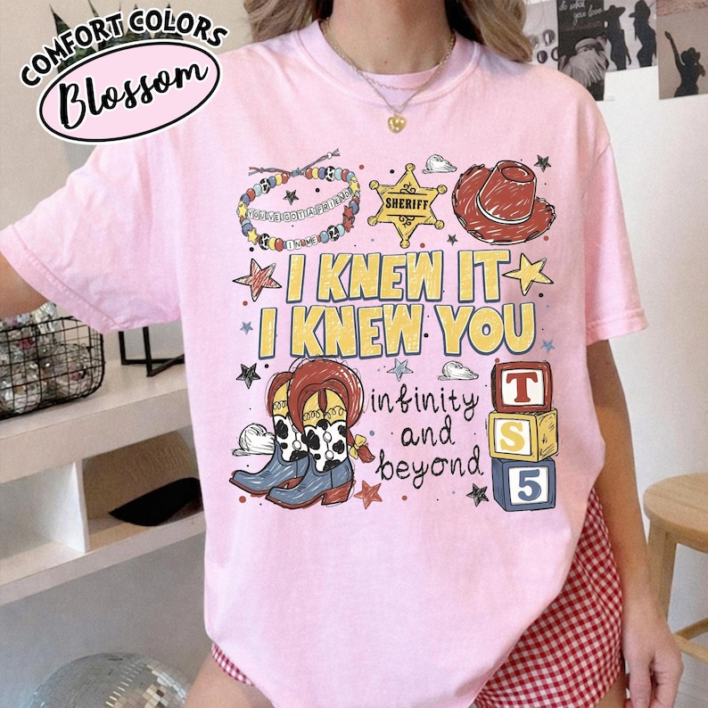 May include: A light pink t-shirt featuring a graphic print with the text I KNEW IT I KNEW YOU infinity and beyond. The design includes a beaded bracelet, a yellow sheriff star, a brown cowboy hat, cow-print boots, and three stacked alphabet blocks.