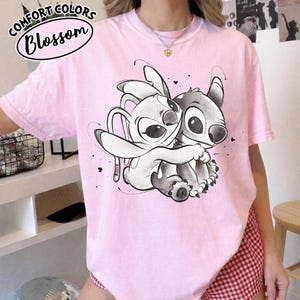 May include: A light pink t-shirt featuring a cartoon illustration of two characters embracing. The characters are in shades of gray and white. The shirt has the words "Comfort Colors" and "Blossom" printed on it.