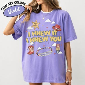 May include: A violet short-sleeve t-shirt featuring a graphic print with the text "I KNEW IT I KNEW YOU" in yellow. The design includes a sheriff badge, a red cowboy hat, cowboy boots, a beaded bracelet with "YOU'VE GOT A FRIEND IN ME" text, and blocks.