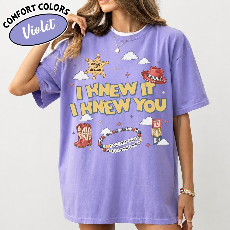 May include: A violet short-sleeve t-shirt featuring a graphic print with the text "I KNEW IT I KNEW YOU" in yellow. The design includes a sheriff badge, a red cowboy hat, cowboy boots, a beaded bracelet with "YOU'VE GOT A FRIEND IN ME" text, and blocks.