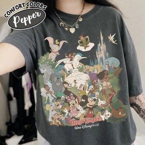 Op de afbeelding: Donkergrijs Comfort Colors T-shirt met een kleurrijke afbeelding van Disney-personages, waaronder Mickey Mouse, Minnie Mouse en Peter Pan. De tekst "Magic Kingdom Walt Disney World" staat eronder. Het shirt heeft een relaxte pasvorm.