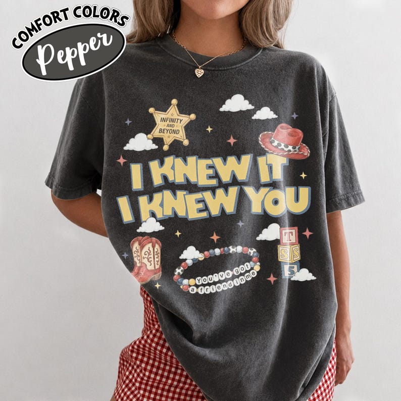 May include: A charcoal gray short-sleeve t-shirt featuring the text I KNEW IT I KNEW YOU in yellow. The graphic includes a sheriff badge with INFINITY AND BEYOND, a red cowboy hat, cowboy boots, a beaded bracelet with YOU'VE GOT A FRIEND IN ME, and blocks.