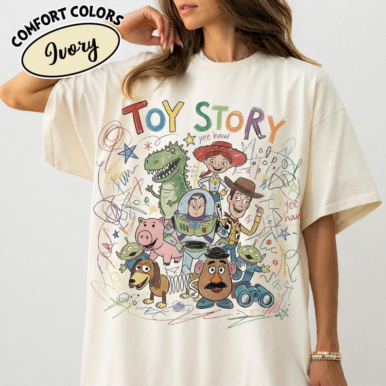 May include: Ivory-colored t-shirt featuring a colorful graphic of Toy Story characters, including Woody, Buzz Lightyear, and Rex. The words "TOY STORY" are written in a playful font above the characters. The shirt also has the words "Comfort Colors" and "Ivory" printed on it.